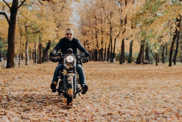 Brutale professionele mannelijke motorrijder rijdt op de fiets draagt zonnebril, handschoenen en een zwarte jas heeft een rit door het herfstpark, een prachtig landschap op de achtergrond met gele bomen en gevallen bladeren rond