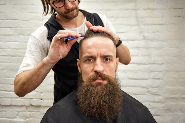 Brute man in de moderne Barber Shop. Kapper maakt van kapsel een man met een lange baard. Kapper doet kapsel met een schaar en kam