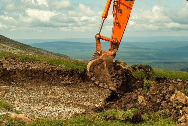 Bucket of a working excavator tears off the grass and soil cover, digging a trench