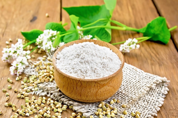 Buckwheat flour from green cereals in a bowl on sacking groats on the table fresh flowers and buckwheat leaves on the background of wooden board