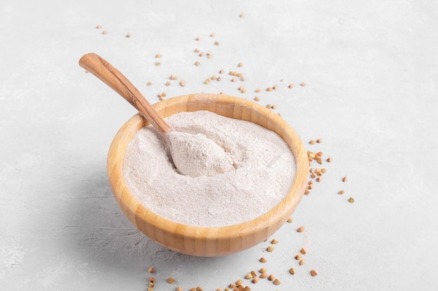 Buckwheat flour in a wooden bowl with wooden spoon and raw green buckwheat grain on grey stone