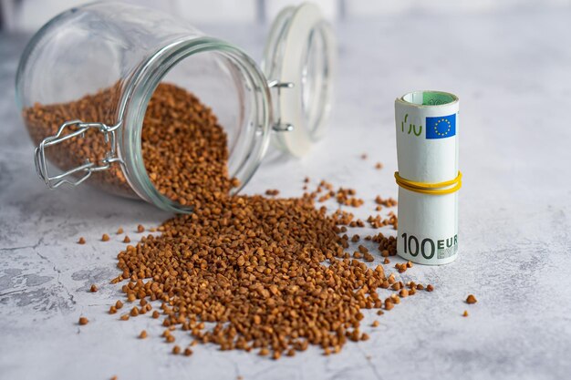 Buckwheat poured from glass jar and stack of money on gray background Inflationfood crisis concept