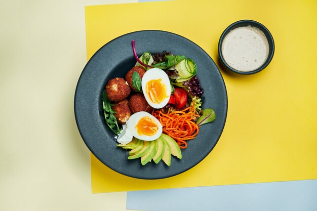 Buddha bowl with chicken croquettes, avcoado, egg, carrot, lettuce and cucumbers in a black plate on a colored surface Close up view. Healthy food