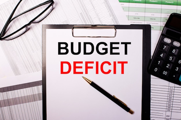 BUDGET DEFICIT is written on a white sheet of paper, near the glasses and the calculator.