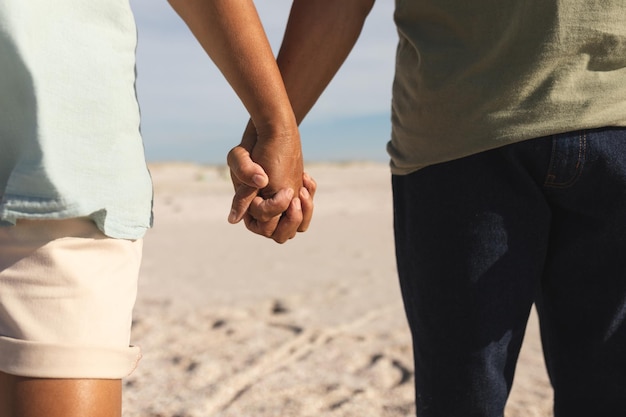 Buik van een multiraciaal senior koppel dat handen vasthoudt en samen op het strand staat op zonnige dag