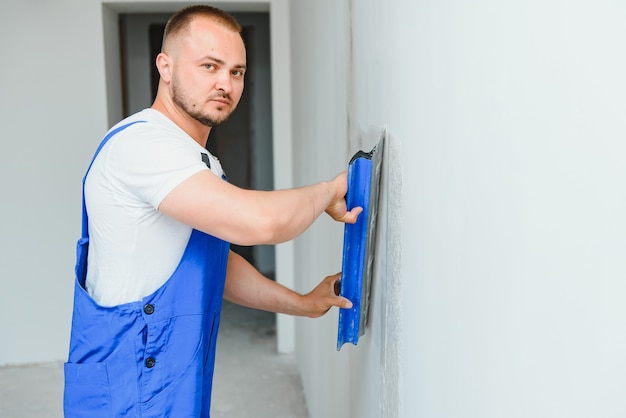 The builder carefully corrects the irregularities of the wall with a trowel. Builder in work clothes against a gray wall. Photo plasterer at work.