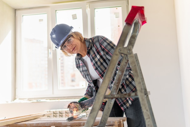 Photo builder cuts grinder pieces of iron. middle-aged woman in a helmet makes repairs in the apartment.