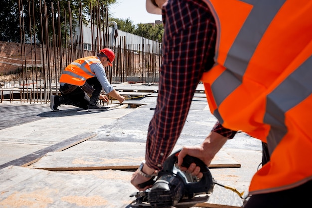 Builder dressed in orange work vest and helmet is using a circular saw on the open building site . .