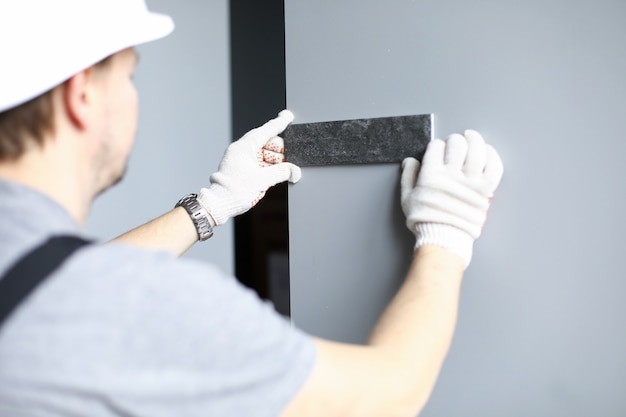 Builder in gloves and helmet picks the color of the tiles for the wall in the apartment. Man applies a sample of building material to the wall