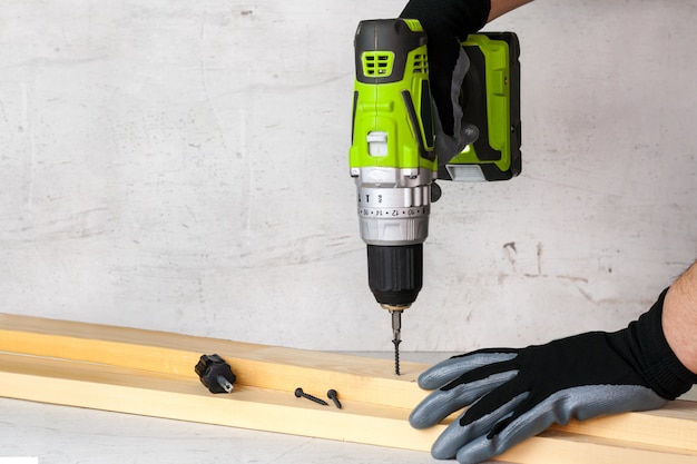 The builder holds in his hand an electric screwdriver on the background of a concrete wall. Screws screw into a wooden beam. DIY