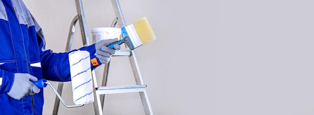 The builder is a painter in a blue suit a brush in his hand and buckets with products for the restoration and painting of the wall on the construction site of the house