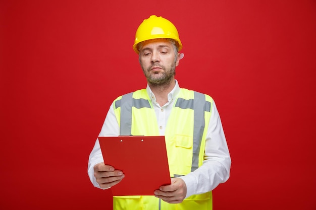 Builder man in construction uniform and safety helmet holding clipboard looking at it with serious face standing over red background