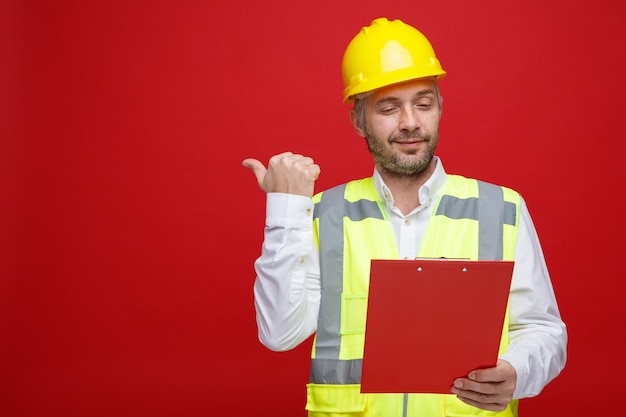 Builder man in construction uniform and safety helmet holding clipboard looking at it with smile on face pointing with thumb to the side standing over red background