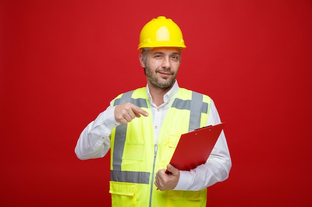 Builder man in construction uniform and safety helmet holding clipboard pointing with index finger at it looking at camera smiling standing over red background