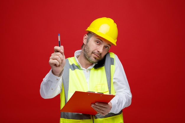 Uomo del costruttore in uniforme da costruzione e casco di sicurezza che tiene appunti parlando sul telefono cellulare che mostra la penna che sembra fiducioso in piedi su sfondo rosso