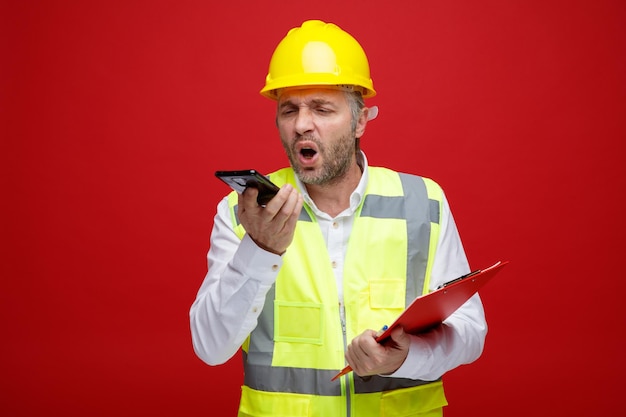 Uomo del costruttore in uniforme da costruzione e casco di sicurezza che tiene appunti parlando sul telefono cellulare urlando arrabbiato e frustrato in piedi su sfondo rosso