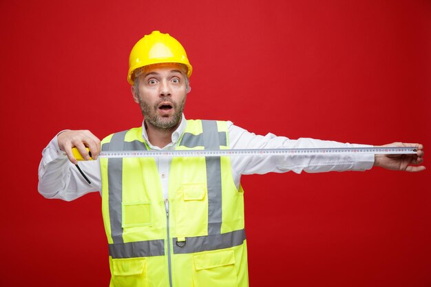 Uomo del costruttore in uniforme da costruzione e casco di sicurezza che tiene nastro di misura guardando la fotocamera stupito e sorpreso in piedi su sfondo rosso