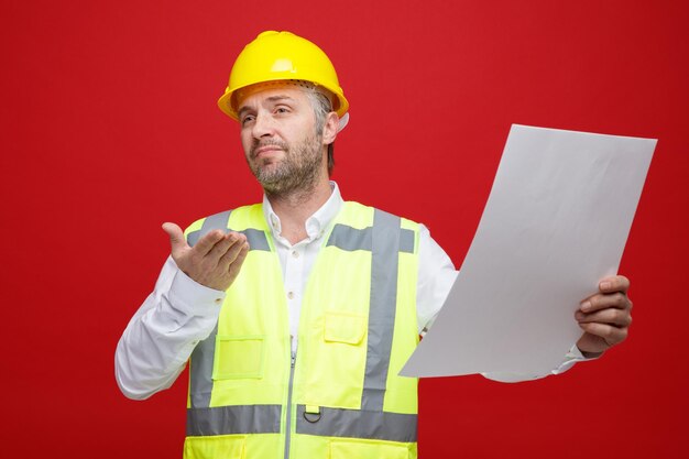 Builder man in construction uniform and safety helmet holding a plan looking confused raising arm in displeasure standing over red background