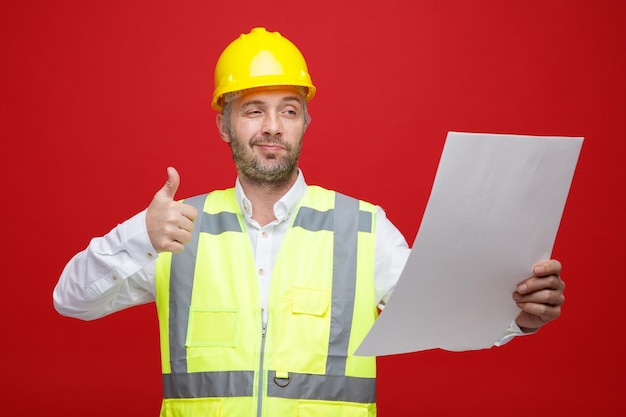 Мужчина-строитель в строительной форме и защитном шлеме держит план, смотрит на него счастливым и позитивно улыбающимся, показывая большой палец вверх, стоя на красном фоне