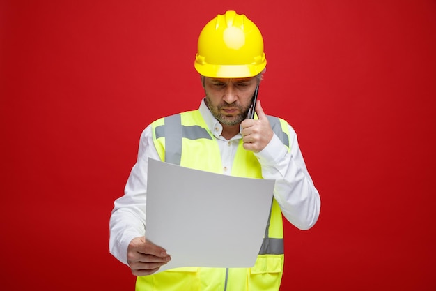 Builder man in construction uniform and safety helmet holding a plan looking at it with serious face talking on mobile phone standing over red background