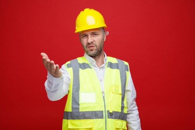 Foto builder uomo in uniforme da costruzione e casco di sicurezza guardando la fotocamera confuso e dispiaciuto alzando il braccio con dispiacere in piedi su sfondo rosso