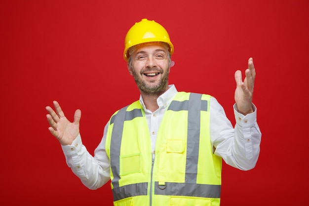 Builder man in construction uniform and safety helmet looking at camera happy and cheerful smiling raising arms standing over red background