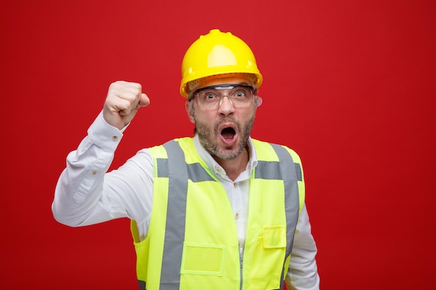Uomo del costruttore in uniforme da costruzione e casco di sicurezza che indossa occhiali di sicurezza pugno di serraggio arrabbiato e frustrato che urla in piedi su sfondo rosso
