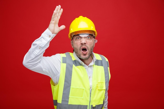 Builder man in construction uniform and safety helmet wearing safety glasses angry and frustrated raising arm in displeasure going wild standing over red background