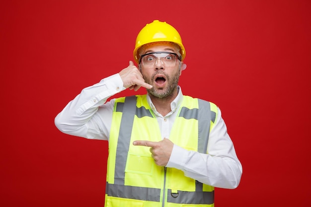 Builder man in construction uniform and safety helmet wearing safety glasses looking at camera amazed making call me gesture pointing with index finger to the side standing red background