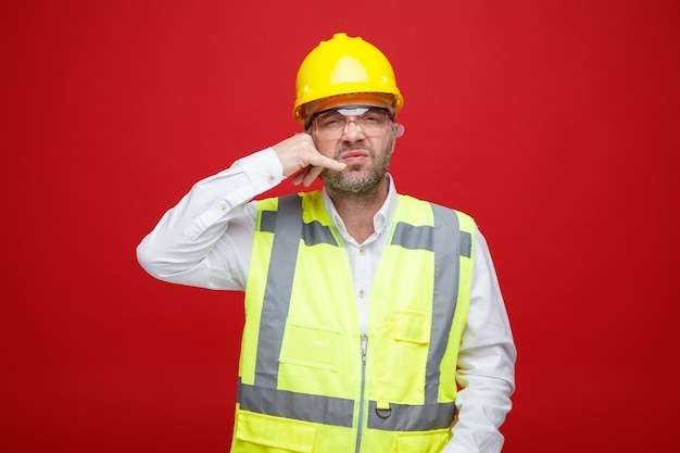 Builder man in construction uniform and safety helmet wearing safety glasses looking at camera displeased making call me gesture standing over red background