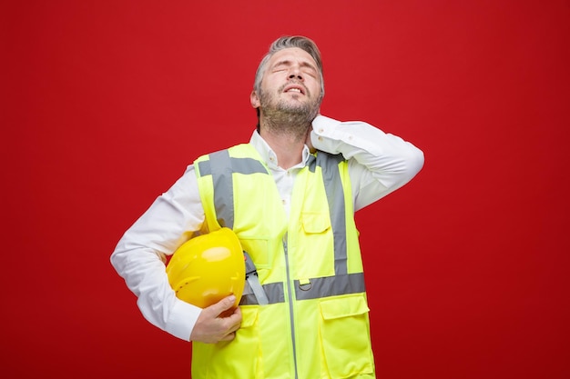 Builder man in construction uniform with safety helmet looking unwell touching his neck feeling pain standing over red background