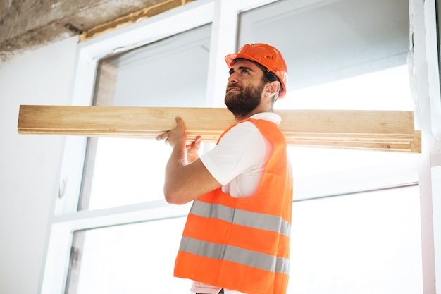 Builder man in hardhat carrying timber on building site close up