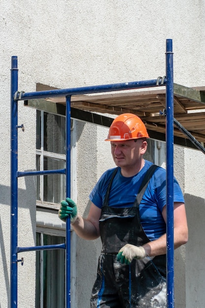 A builder in an orange helmet works on scaffolding repair of\
the facade of the building wall painting plastering and wall\
insulation construction site during the repair of the walls of the\
house