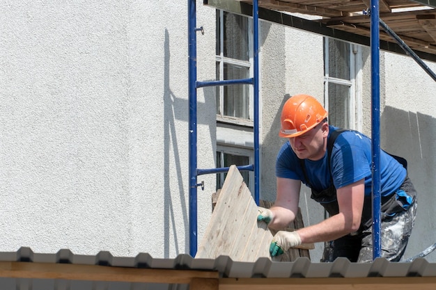 A builder in an orange helmet works on scaffolding Repair of the facade of the building wall painting plastering and wall insulation Construction site during the repair of the walls of the house