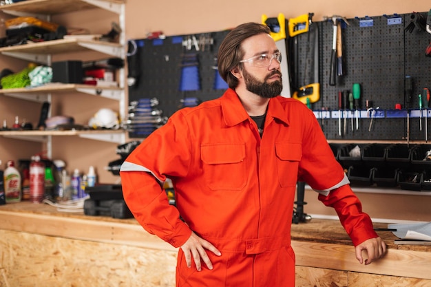 Builder in orange work clothes and protective eyewear thoughtfully looking aside with tools on background in workshop