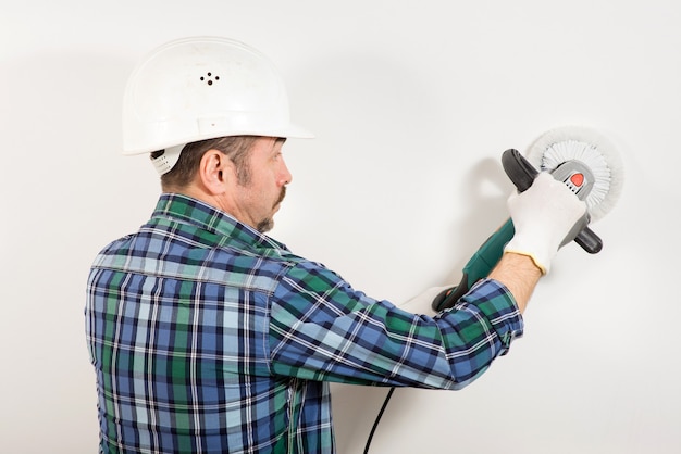 A builder in a safety helmet is grinding a white wall with a grinder