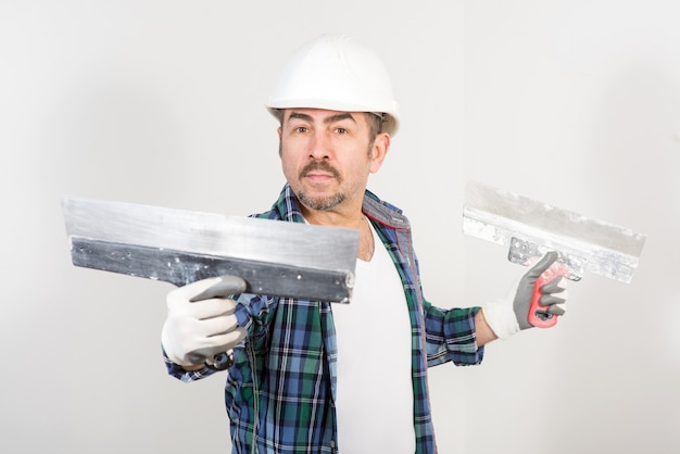 Builder in safety helmet with two putty knifes on white wall