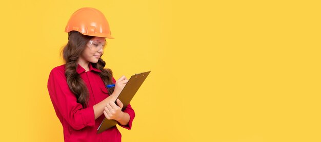 Builder teenager girl in helmet smiling kid girl in safety glasses and helmet hold folder for making notes mark data child builder in helmet horizontal poster design banner header copy space