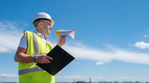 Builder in a white helmet shouts into a megaphone against a blue sky background