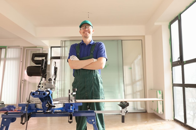 Builder with crossed arms stands by disk saw machine in repaired room smiling worker with