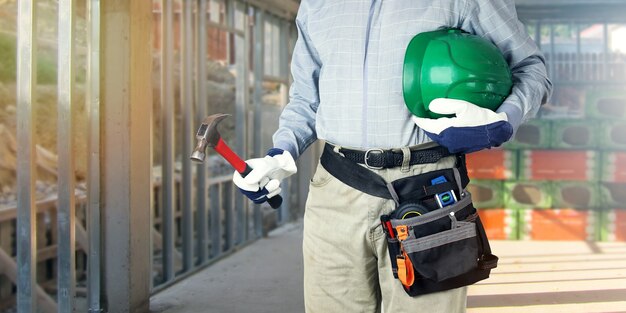 Builder with tools holds hammer and hard hat in hands