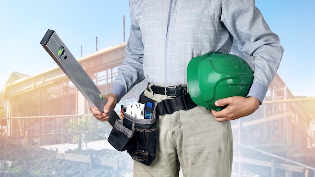 Builder with tools holds level and hard hat on background of building construction