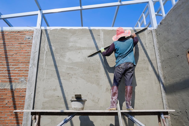 Builder worker on platform using long trowel to polishing concrete wall in house construction site