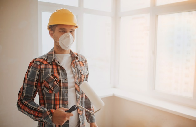 Il costruttore lavora in cantiere e misura il soffitto. un lavoratore in un casco arancione e un rullo di vernice dipinge il muro.