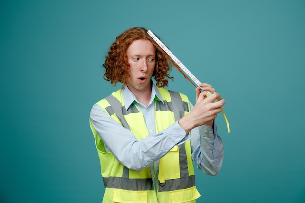 Builder young man in construction uniform holding tape measure looking at it amazed and surprised standing over blue background