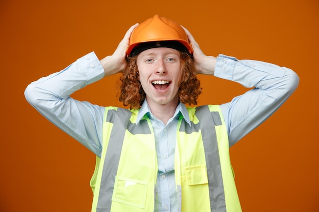 Giovane costruttore in uniforme da costruzione e casco di sicurezza felice ed eccitato tenendosi per mano dietro la testa sorridendo ampiamente in piedi su sfondo arancione