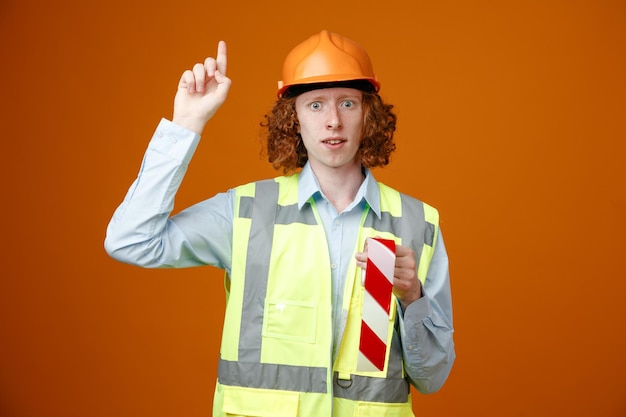 Builder young man in construction uniform and safety helmet holding adhesive tape looking at camera worried showing index finger warning standing over orange background