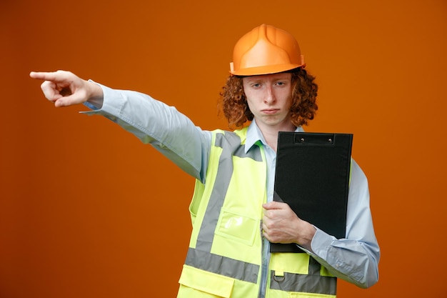 Builder young man in construction uniform and safety helmet holding clipboard looking at camera with serious face pointing with index finger at something standing over orange background