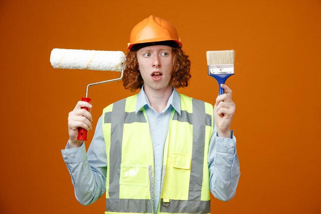 Giovane costruttore in uniforme da costruzione e casco di sicurezza che tiene rullo di vernice e pennello che sembra confuso avendo dubbi in piedi su sfondo arancione