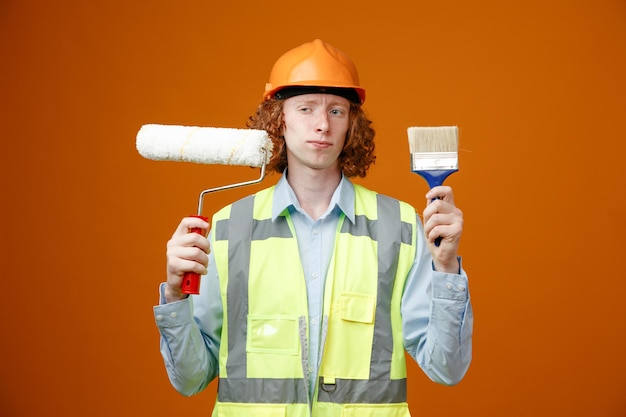 Builder young man in construction uniform and safety helmet holding paint roller and brush looking confused making choice standing over orange background